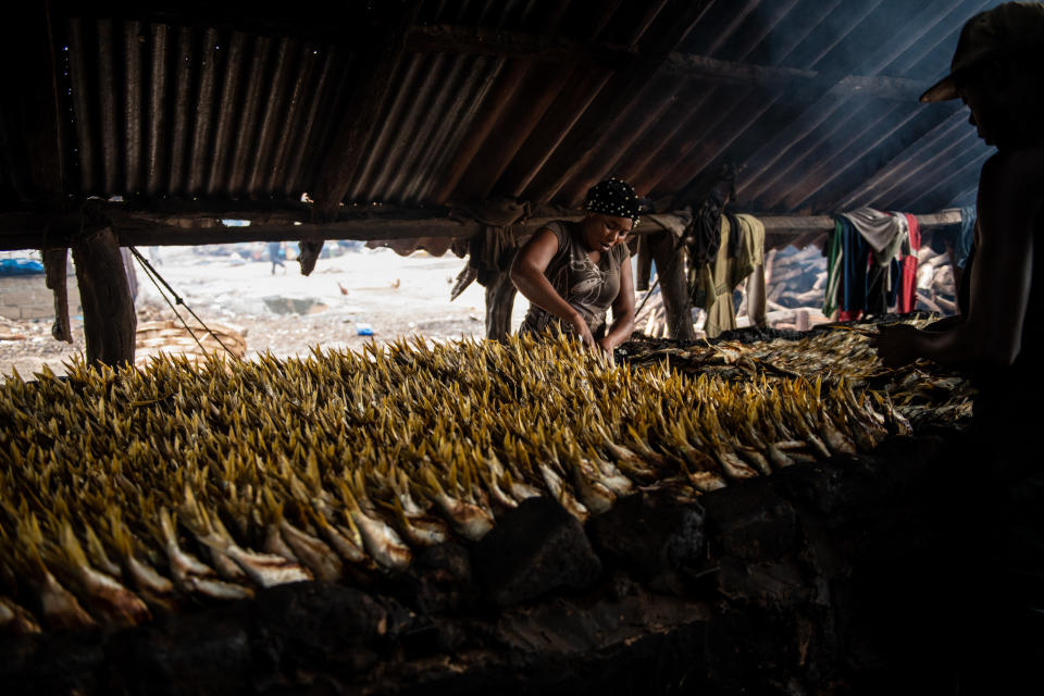 Wide shot of a woman and a man carefully sorting dead fish under a roof with the shore in view.