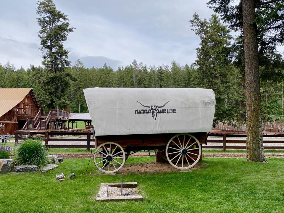 A covered wagon parked on a green lawn in front of trees and a wood fence.