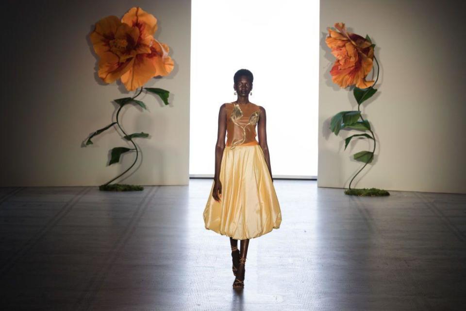 A model walks down a runway wearing a dress with a flower motif. The walls behind her also have two large flower motifs.