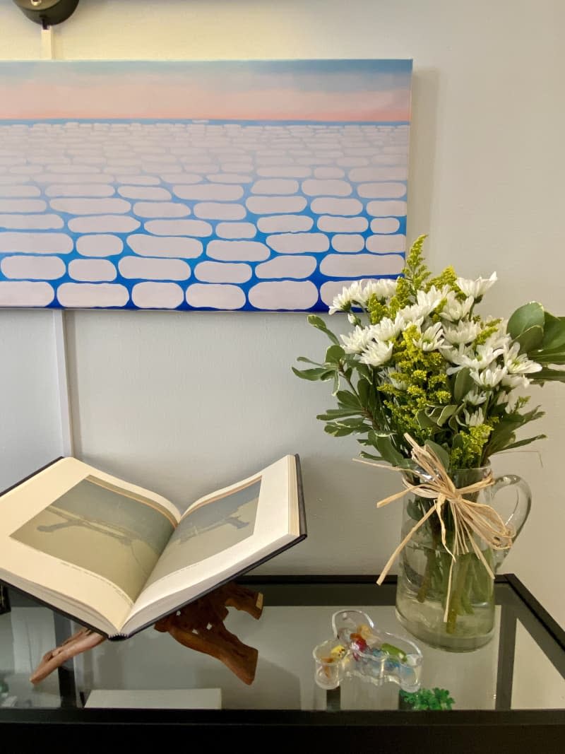 A piece of art above a book that is on display next to a vase full of flowers.