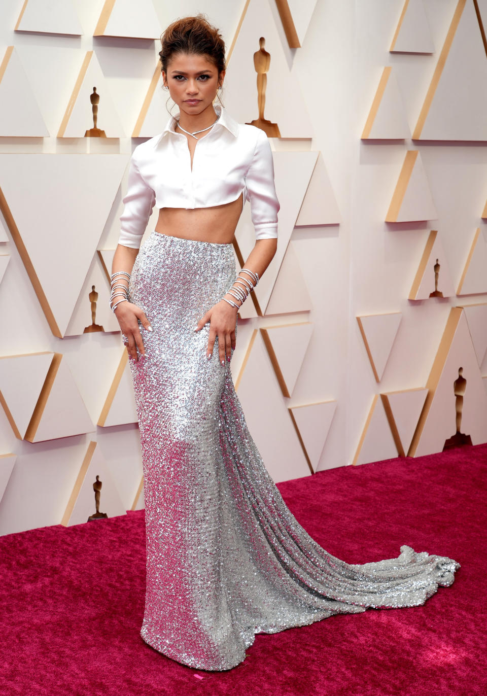 Zendaya attends the 94th Annual Academy Awards at Hollywood. She wears a cropped white blouse which exposes her tummy, with a long silver sequinned floor length skirt with a train. She wears silver bangles up her arms.