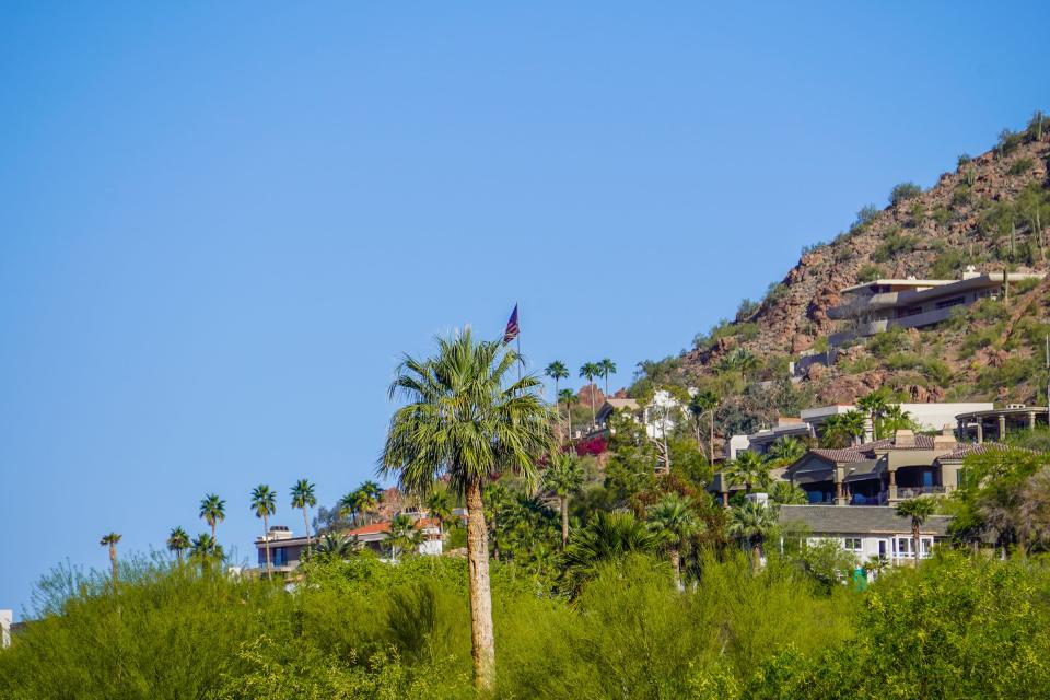 Mansion on the side of a red mountain dotted with cacti. Blue, clear skies in the background and palm trees in front