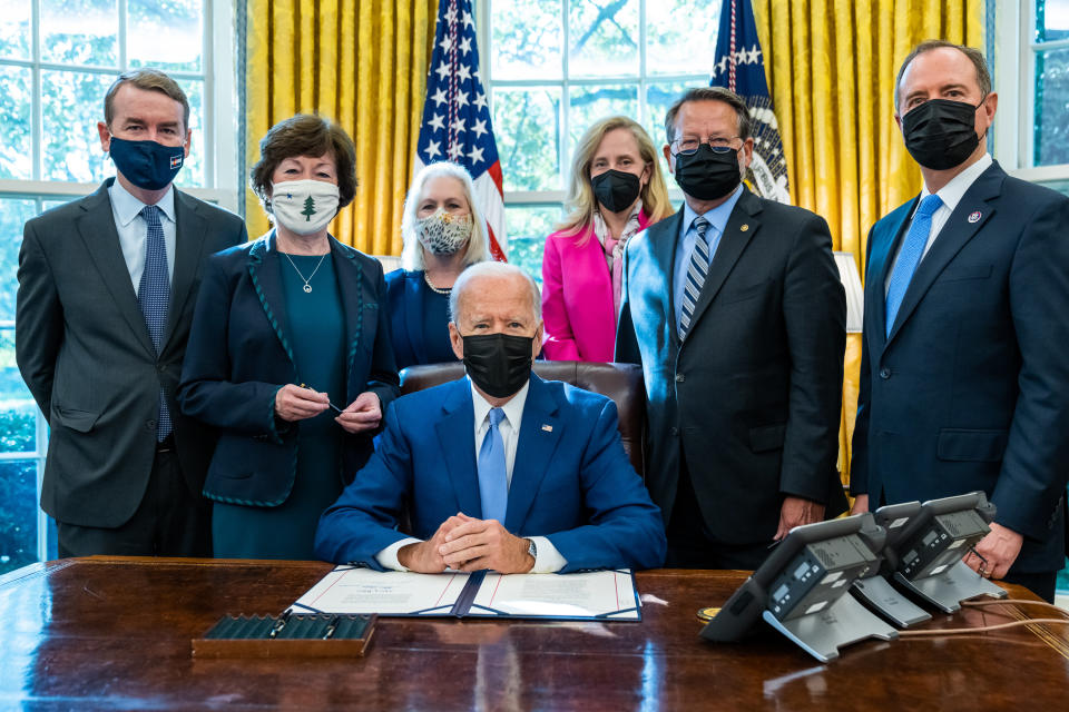 President Biden sits behind the desk in the Oval Office with officials standing behind him, after signing a bill into law.