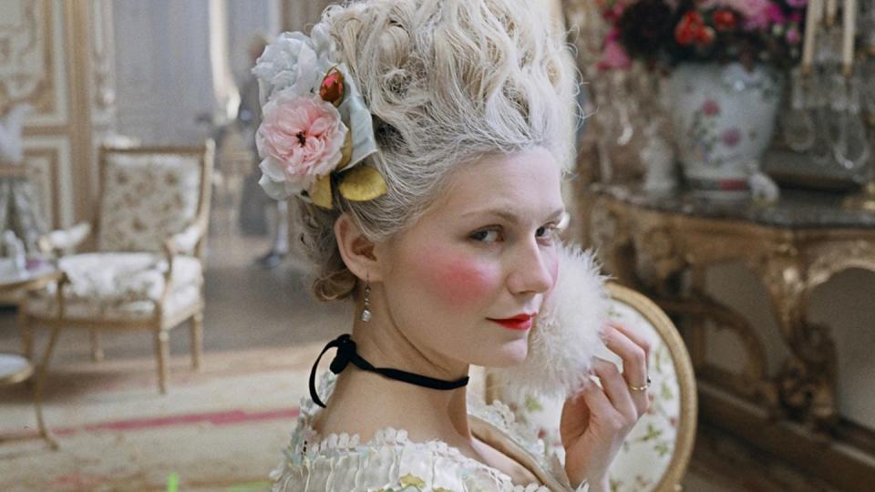 kirsten dunst as marie antoinette sits in a lavish room with a large pink floral bouquet and furniture behind her, she wears a lacy dress, black necklace and flowers in her hair, she holds a white puff to her face as she eyes the camera