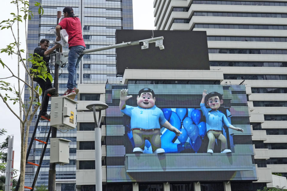 Workers install a traffic CCTV pole as a an LED board is seen featuring a video of Indonesian presidential candidate Prabowo Subianto, left, and his running mate Gibran Rakabuming Raka as animation characters, at the main business district in Jakarta, Indonesia, Tuesday, Feb. 6, 2024. As Indonesia votes this month to replace popular President Joko Widodo, all three candidates have all been aggressively seeking to win the votes of younger people, reaching out to them on the apps they use, through the K-pop music many love, and even video gaming events. (AP Photo/Achmad Ibrahim)