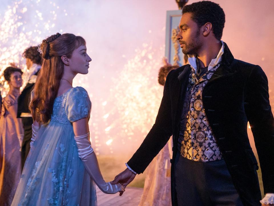 daphne and simon dancing in bridgerton. daphne is wearing a sheer blue gown, her hair worn half up, and simon is wearing a shining waistcoast and black overcoat. they are looking intently into each others eyes