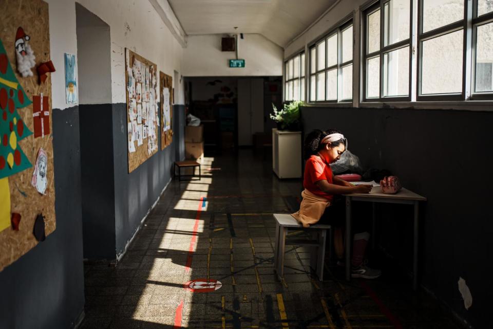 A young girl seated at a desk in a hallway under a row of windows