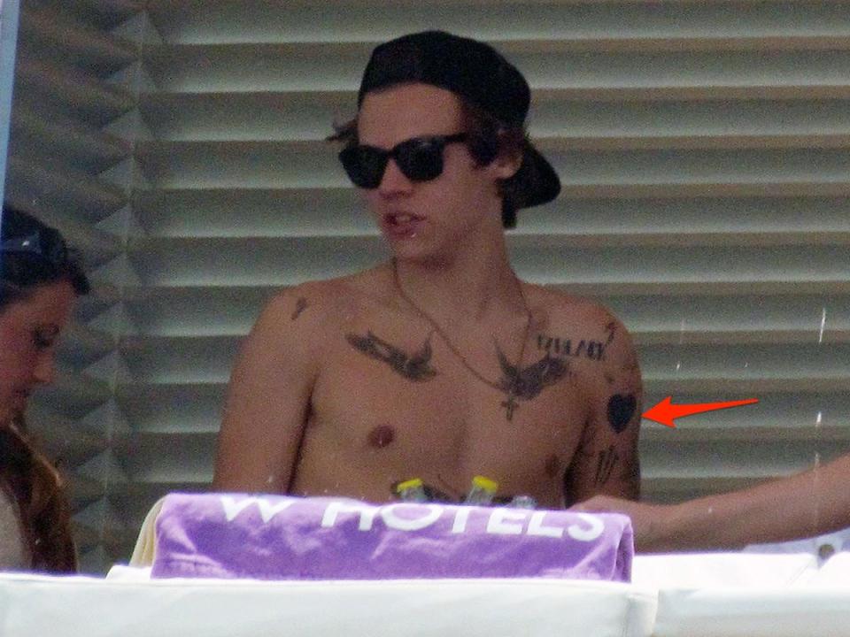 A red arrow pointing to a heart tattoo on Harry Styles' left arm.