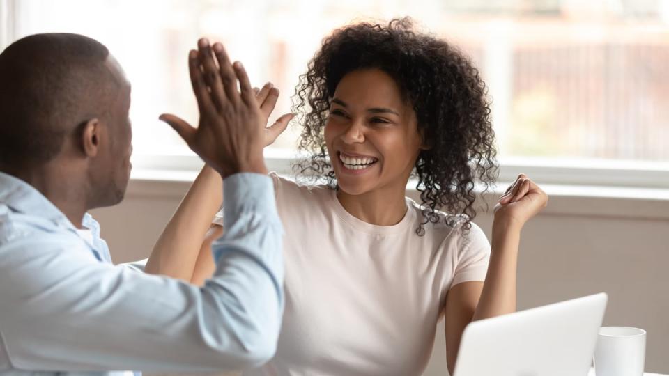 Two people high five while sitting at a table with a laptop and a mug. 