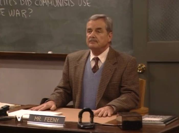 Mr. Feeny sits at his desk in a classroom, wearing a blazer over a sweater vest. His nameplate reads "Mr. Feeny." Books and a chalkboard are visible in the background