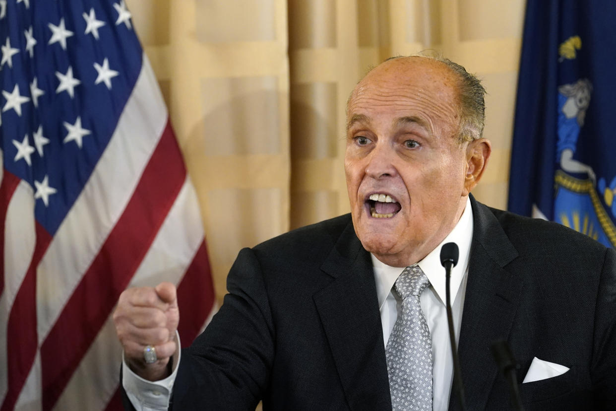 Rudy Giuliani next to an American flag speaking at a microphone with a raised fist.