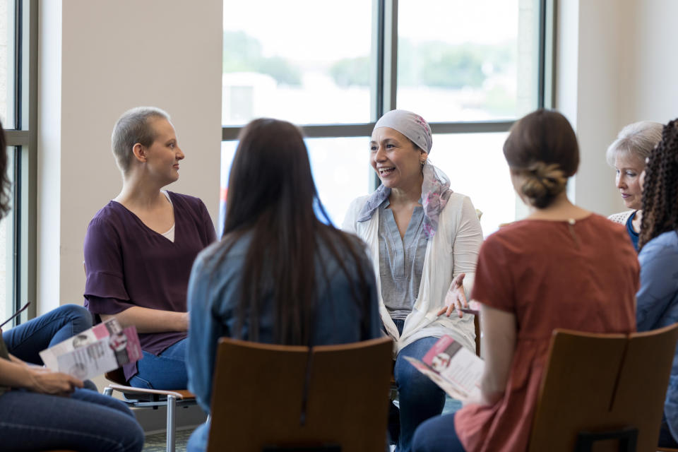 A group of women, including two with headscarves, sit in a circle, engaged in conversation, holding pamphlets