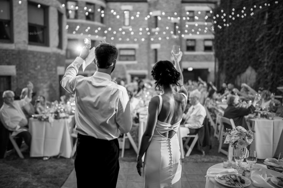 A black and white photo of a bride and groom raising a glass to their guests at their wedding reception.