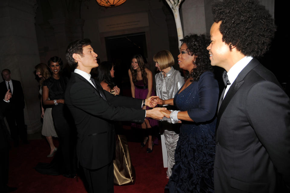 Oz and Oprah at a black-tie event, holding each others' hands at a distance and surrounded by others dressed formally at an indoor venue