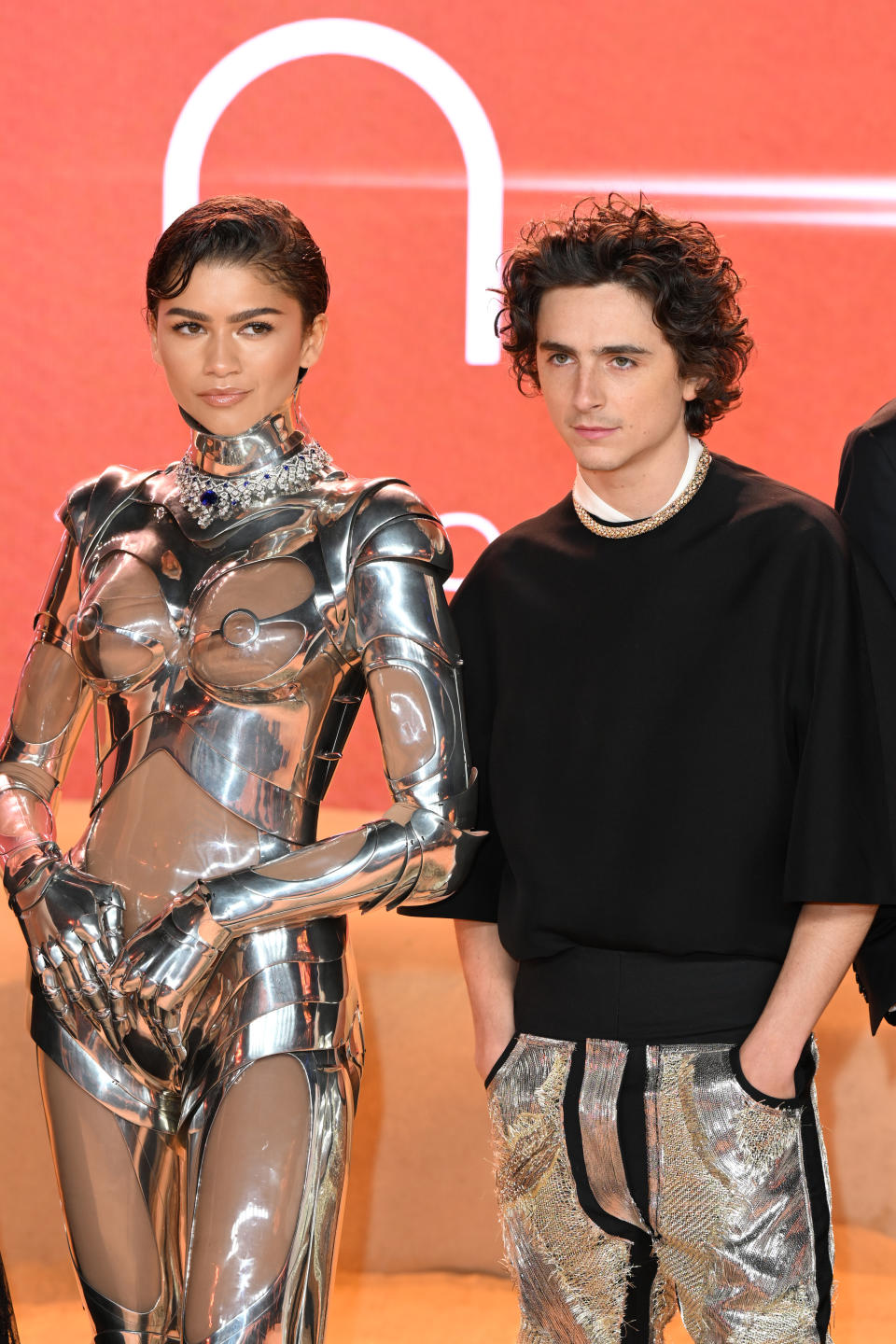 Two individuals on the red carpet. Person on the left in a metallic structured outfit. Person on the right in a black top and shimmery trousers