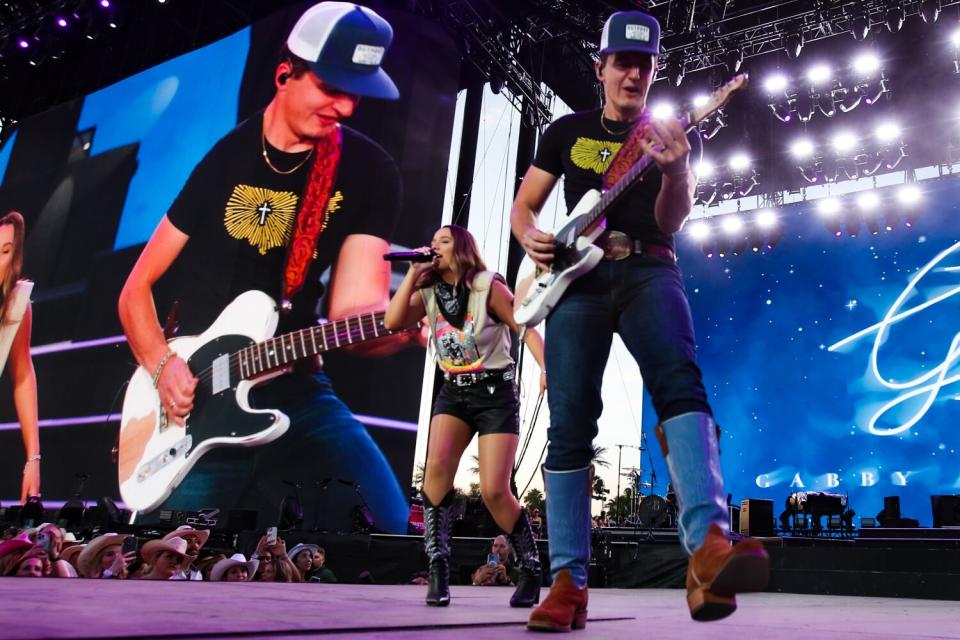 A woman sings and a man plays guitar on a stage with a huge screen showing them perform.