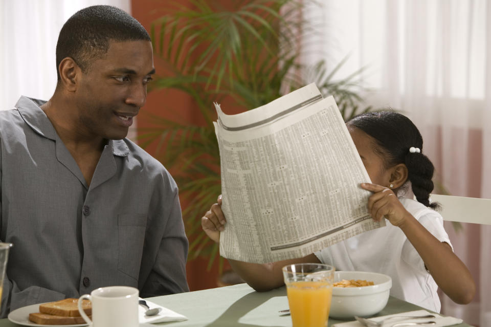 A man in pajamas, sitting at a table with toast, smiles at a little girl holding up a newspaper while eating cereal. A glass of orange juice is on the table