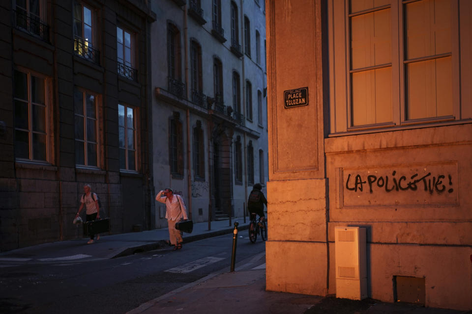 People pass by a wall illuminated by a burning barricade on which is written 'Police kills' in the center of Lyon, central France, Friday, June 30, 2023. French President Emmanuel Macron urged parents Friday to keep teenagers at home and proposed restrictions on social media to quell rioting spreading across France over the fatal police shooting of a 17-year-old driver. Writing on wall reads in French "Justice for Nahel" (AP Photo/Laurent Cipriani)