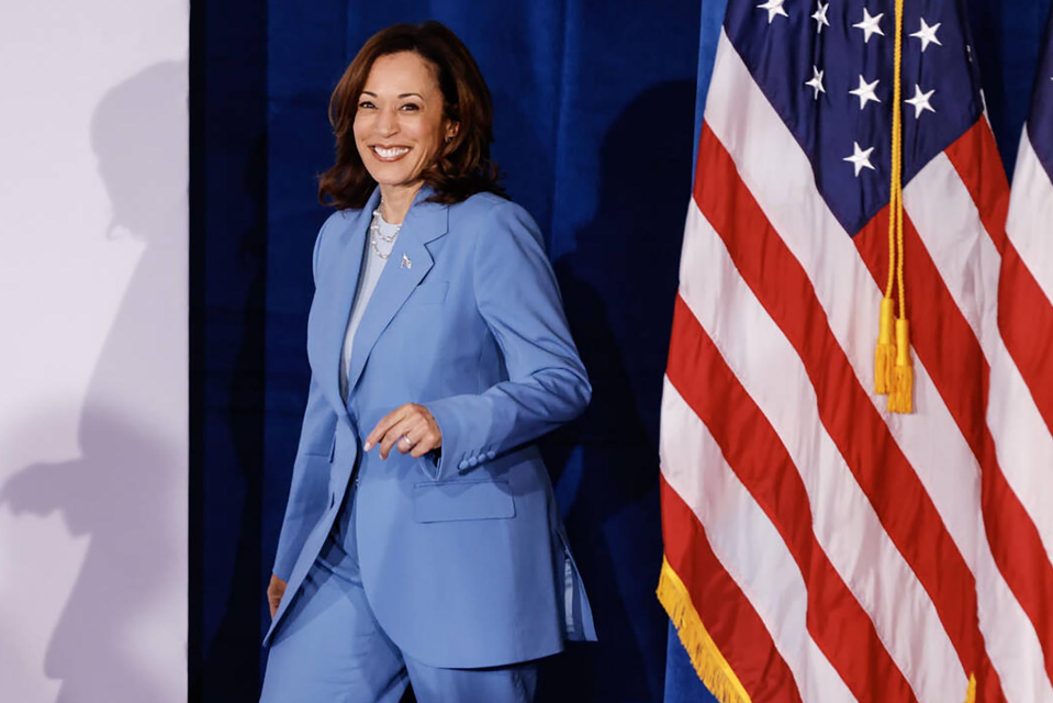 Kamala Harris in a blue suit smiles while walking past an American flag
