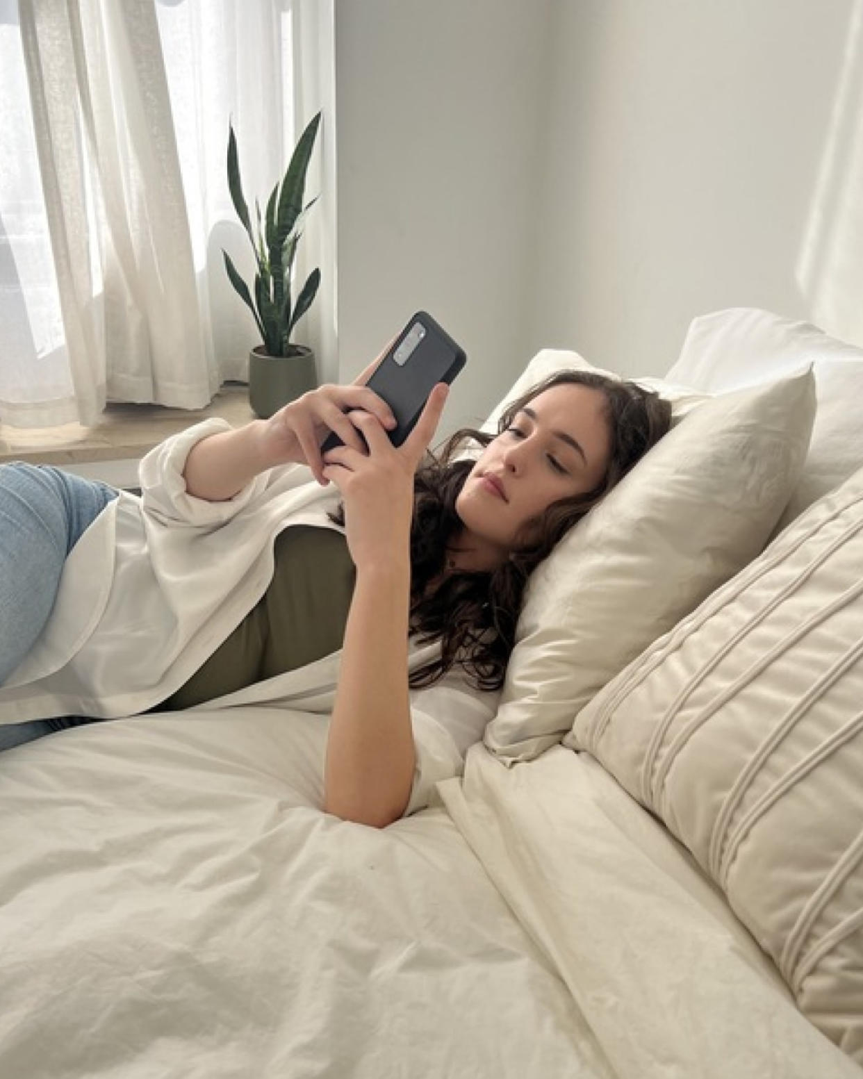 Woman lays on her bed with her head on a Blissy Silk Pillowcase as she scrolls on her phone (Courtesy Mili Godio)