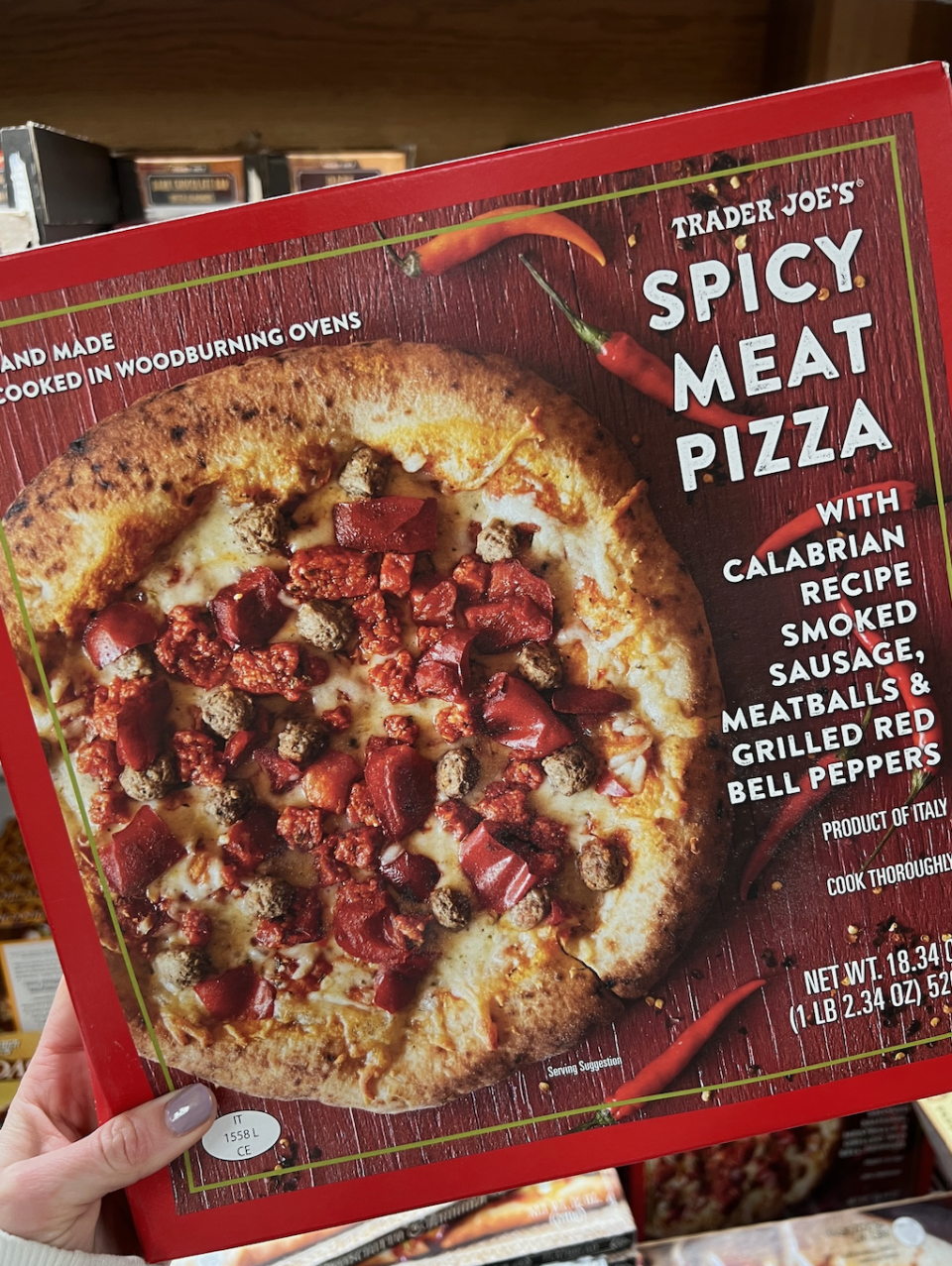 Person holding a Trader Joe's Spicy Meat Pizza box showing pizza with Calabrian chili sauce, smoked sausage, meatballs, and red bell peppers