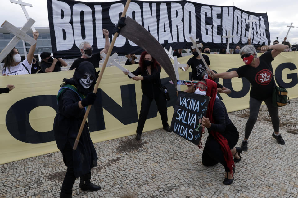 A demonstrator holds the Portuguese message "Vaccination saves lives", below, during a performance with a protester dressed as death and wearing a mock presidential sash, during a protest against President Jair Bolsonaro's handling of the COVID-19 pandemic outside Planalto presidential palace in Brasilia, Brazil, Friday, March 19, 2021. The banner at top reads "Bolsonaro genocide." (AP Photo/Eraldo Peres)