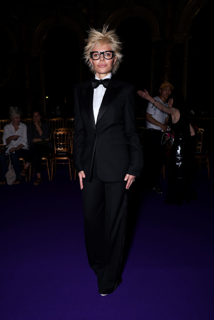 Lisa Rinna dressed in a black tuxedo with a bow tie and large glasses, standing on a purple carpet at an event