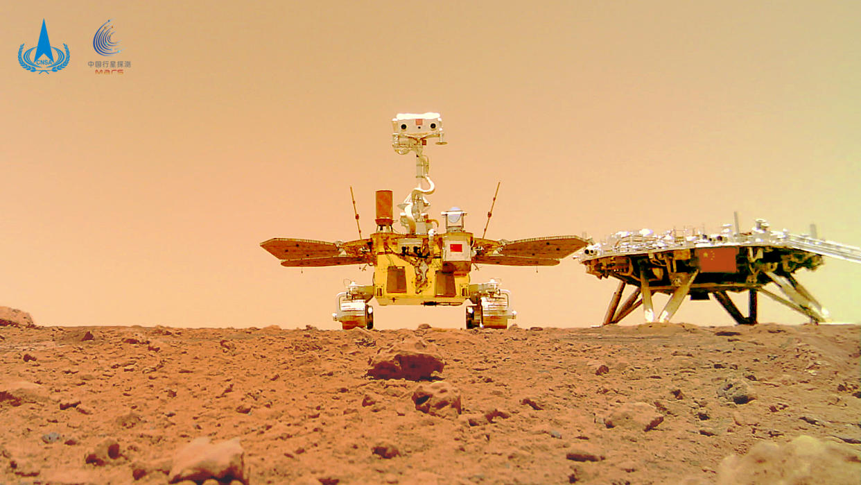  A golden rover and dark-colored lander sit on the red martian surface, with a dusty reddish-brown sky in the background. 