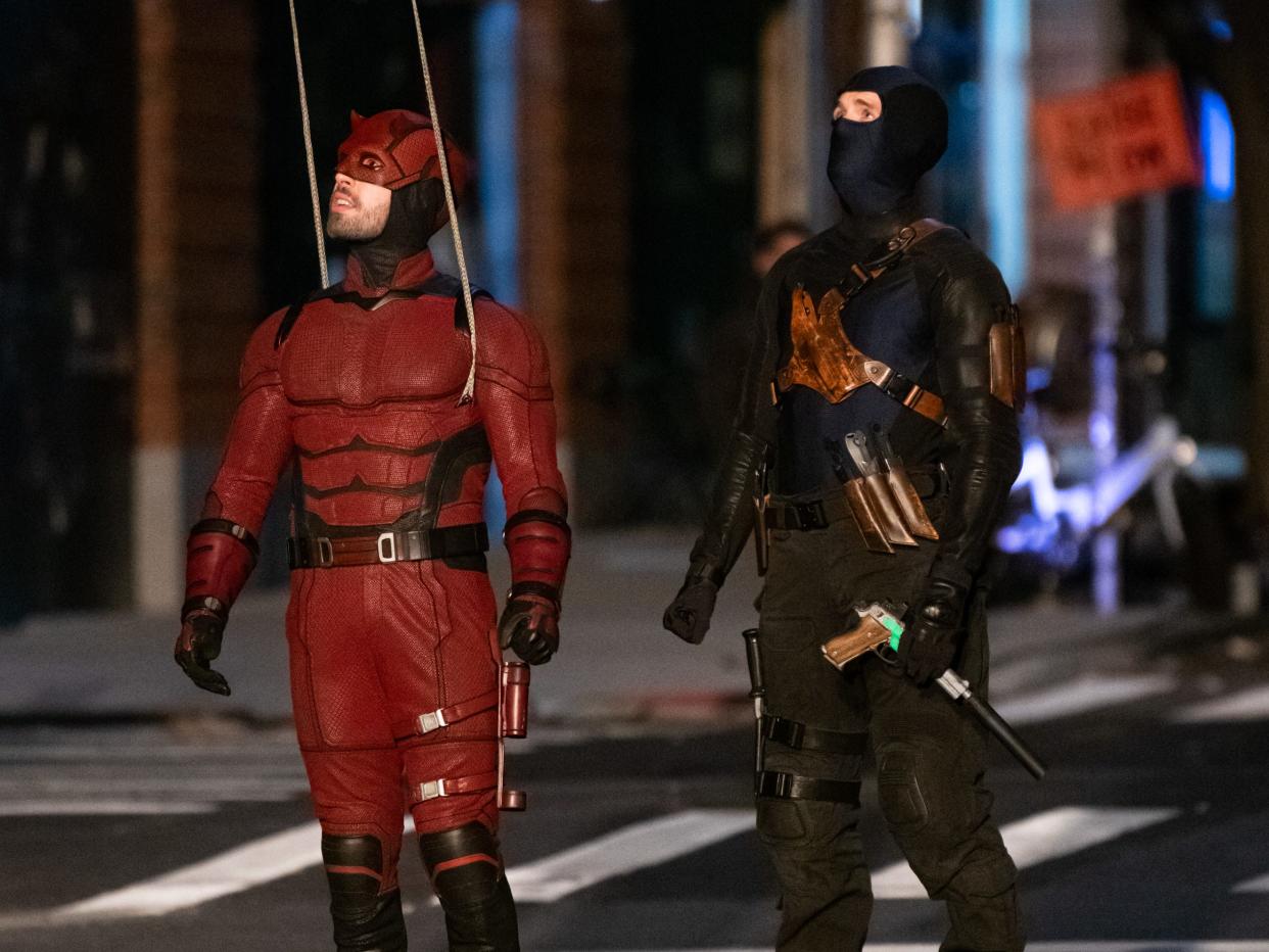 A man in a red suit with black detailing, black boots, and a red mask with horns on it attached to two wires. He's standing next to another man holding a pistol in a dark blue balaclava and a black outfit with a leather harness.