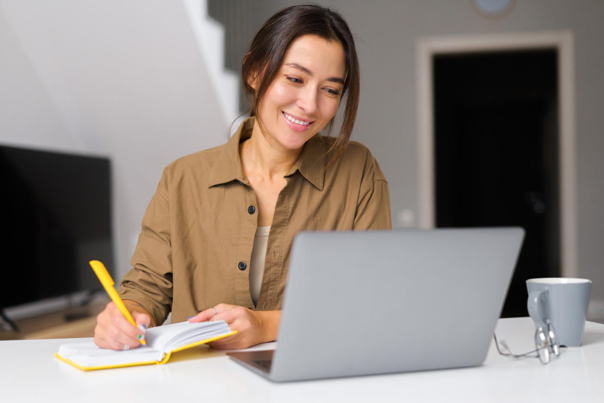 Smiling kind young woman works from home. She sitting at the kitchen table In front of the laptop and notebook. Attractive girl