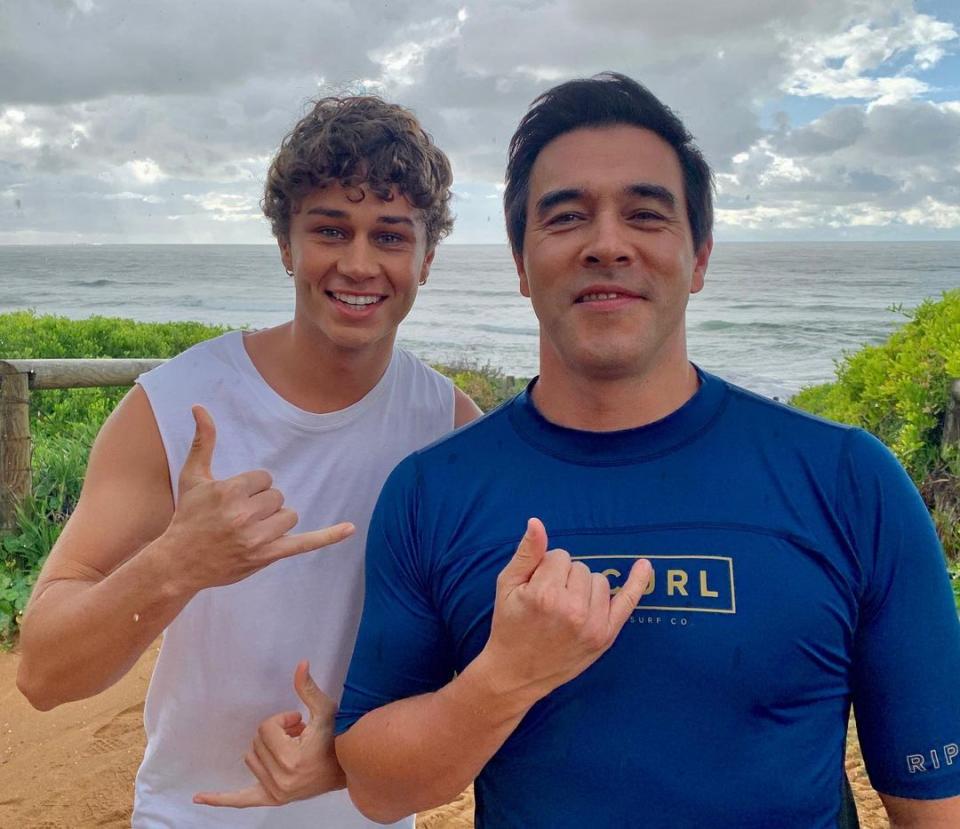 James Stewart in a blue rash shirt and Matt Evans in a white singlet pose for the camera, making surfing hand gestures