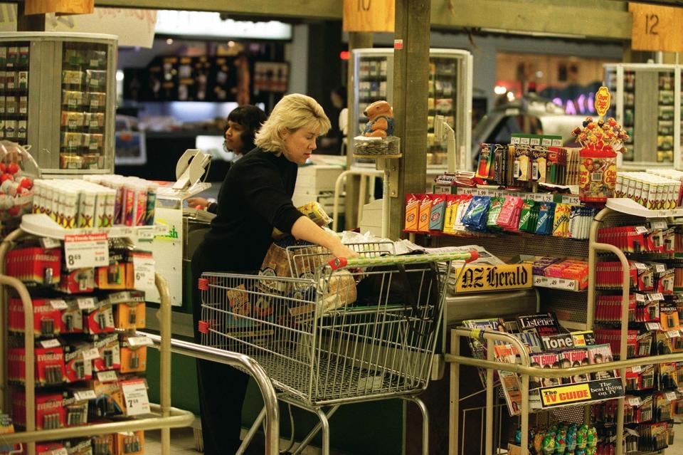 A woman pushes a shopping cart filled with groceries through a store aisle. Shelves are stocked with various snacks and other items