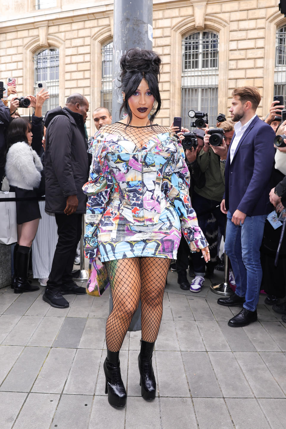 Cardi B in a printed dress, fishnet stockings, and boots, posing for photos while surrounded by fans and paparazzi on a city street