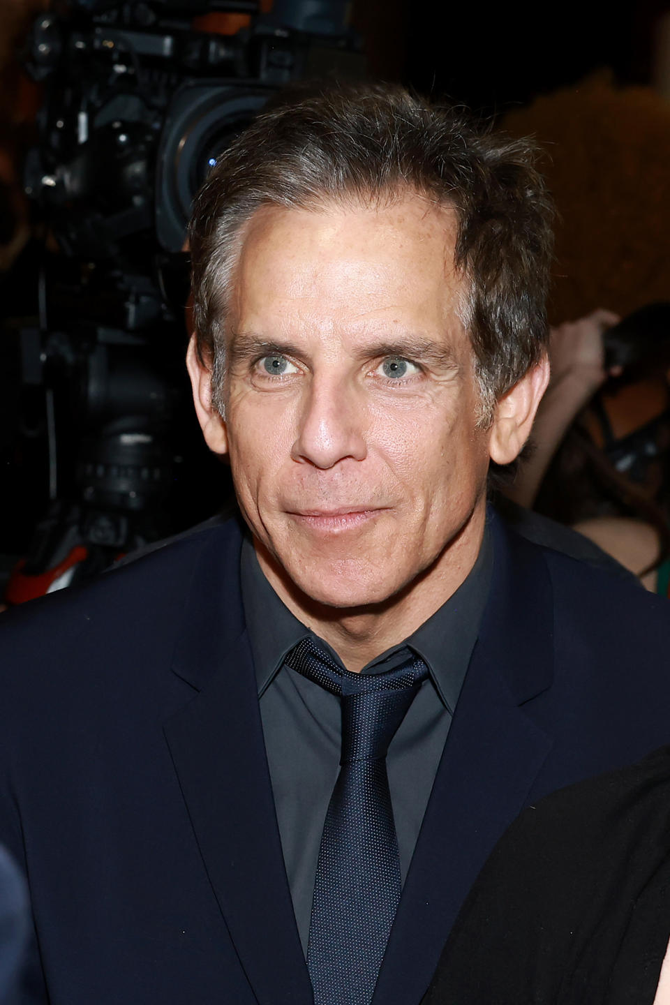 Ben Stiller, wearing a dark suit and tie, attends an event with cameras in the background