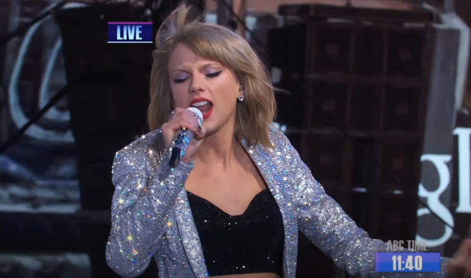 Taylor Swift performing live, wearing a glittery outfit, singing into a microphone
