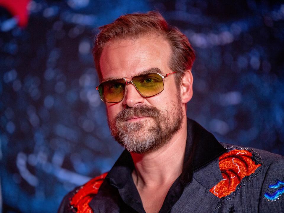 david harbour looking to the side of the camera, wearing yellow-tinted sunglasses, with a trimmed beard, and an embroidered suit jacket