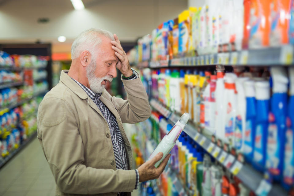 Elderly man with a concerned expression inspects a product in a supermarket aisle filled with household items