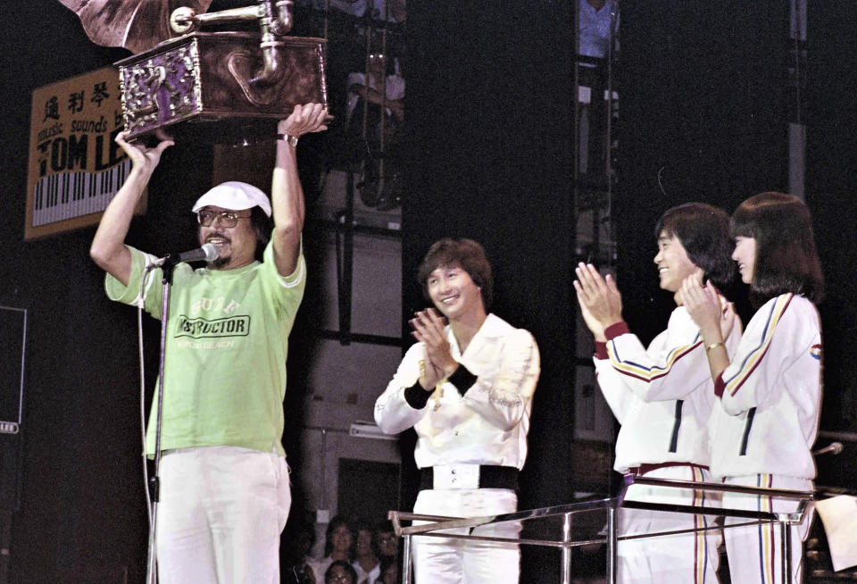 In this undated photo released by Radio Television Hong Kong (RTHK), Hong Kong DJ, Ray Cordeiro, also known as Uncle Ray, left, receives a souvenir presented by Hong Kong singers Sam Hui, second left, and Albert Au, second right, as he bid farewell to the civil service in 1980 on behalf of RTHK in a concert. After more than seven decades in radio, the 96-year-old Hong Kong DJ bid farewell to his listeners Saturday, May 15, 2021 with “Time to Say Goodbye,” sung by Sarah Brightman and Andrea Bocelli. (Radio Television Hong Kong via AP)