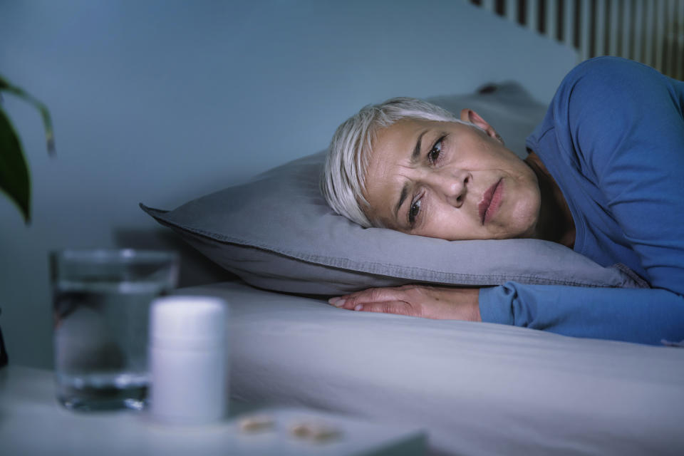 A woman with short gray hair lies in bed looking thoughtful, with a glass of water and a pill bottle on the bedside table
