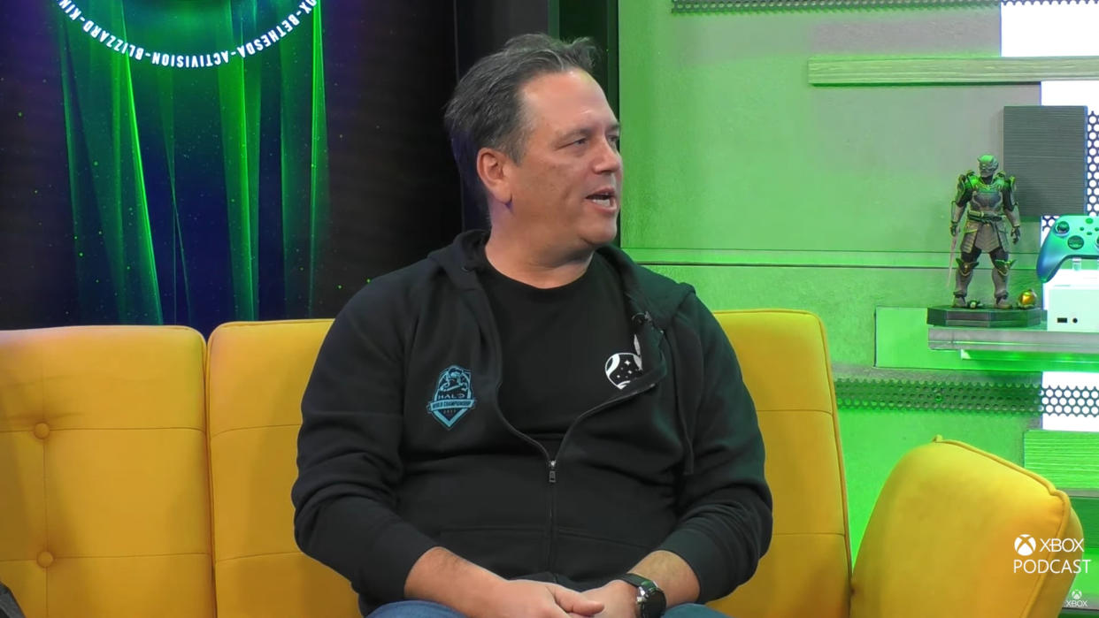  Phil Spencer sitting on a yellow couch with a green and blue Xbox background behind him. 