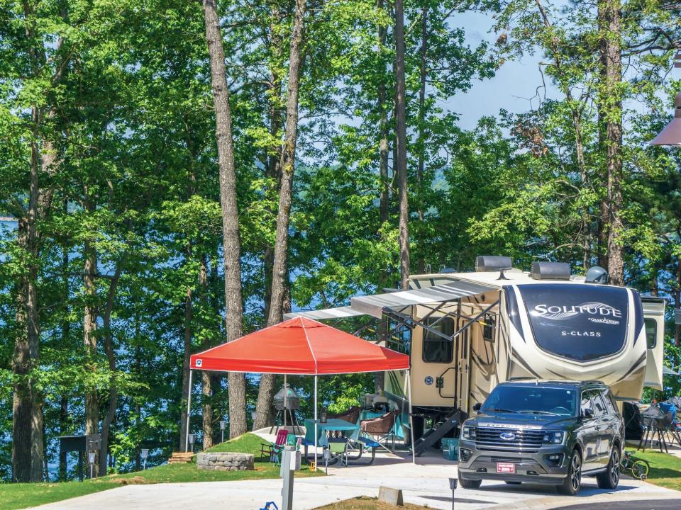 A RV and SUV parked next to outdoor lounge chairs and shade among trees