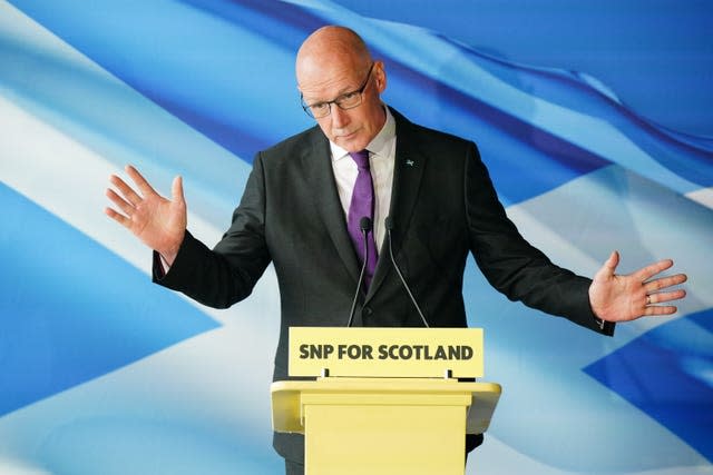 John Swinney, with hands raised, speaking from an SNP lectern, with a Saltire flag backdrop