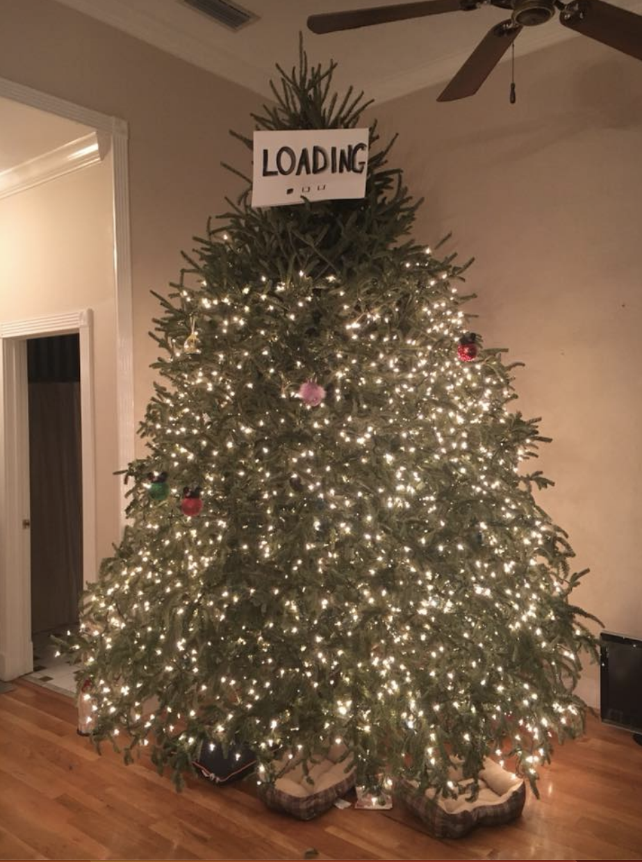 A Christmas tree is covered in lights until about 80% of the way up, where the lights stop and there is no tree topper, only a paper sign saying "loading"