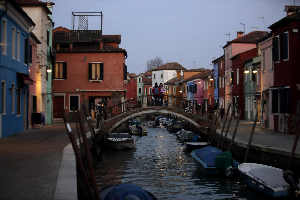 In this image taken on Thursday, Jan. 16, 2020, people stand on a bridge at the Burano island, Italy. The Venetian island of Burano's legacy as a fishing village remains the source of its charms: the small colorful fishermen's cottages, traditional butter cookies that were the fishermen's sustenance at sea and delicate lace still stitched by women in their homes. As the island's population dwindles, echoing that of Venice itself, so too are the numbers of skilled artisans and tradespeople who have kept the traditions and economy alive. (AP Photo/Luca Bruno)