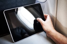 An ipad, propped on the folding table within an aeroplane, areoplane window is reflected on the tablet screen