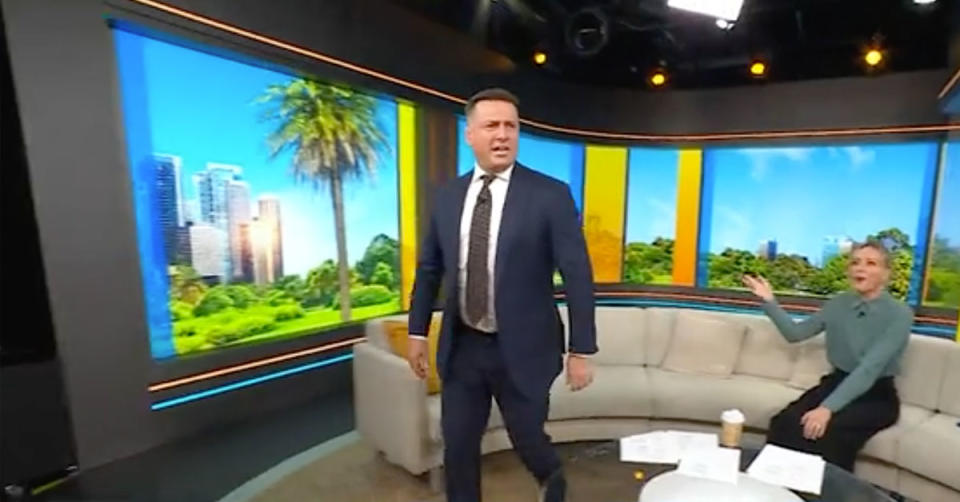 Karl Stefanovic wears a navy suit and black tie, he is walking off the set of the Today show while host Allison Langdon sits on the couch wearing a green skivvy and black skirt.