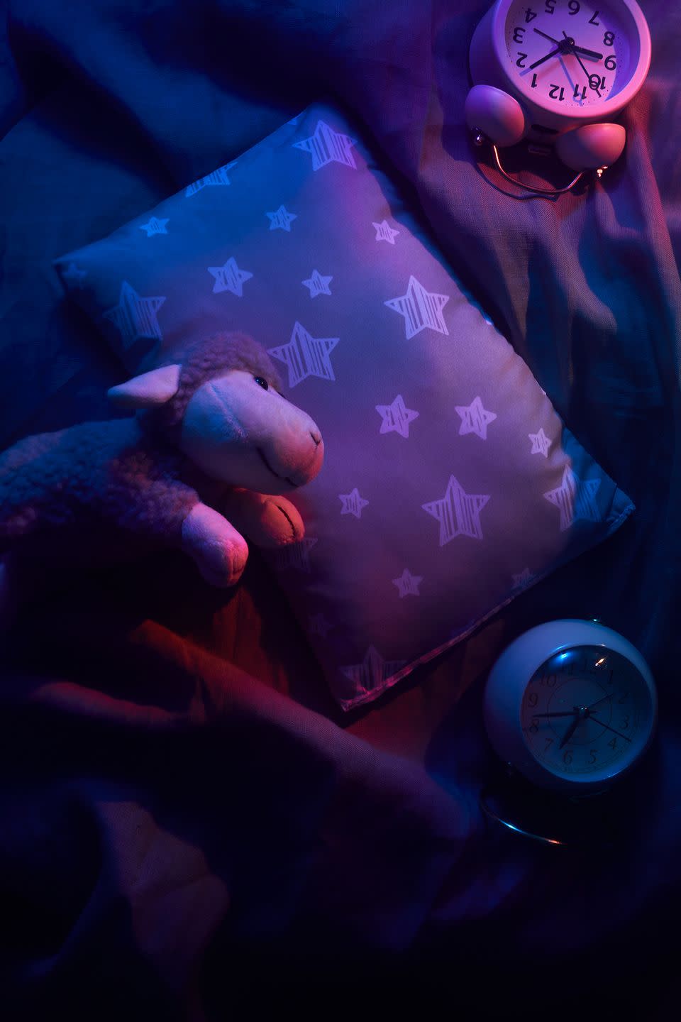 a stuffed animal sheep lying on a bed with a clock and a blanket