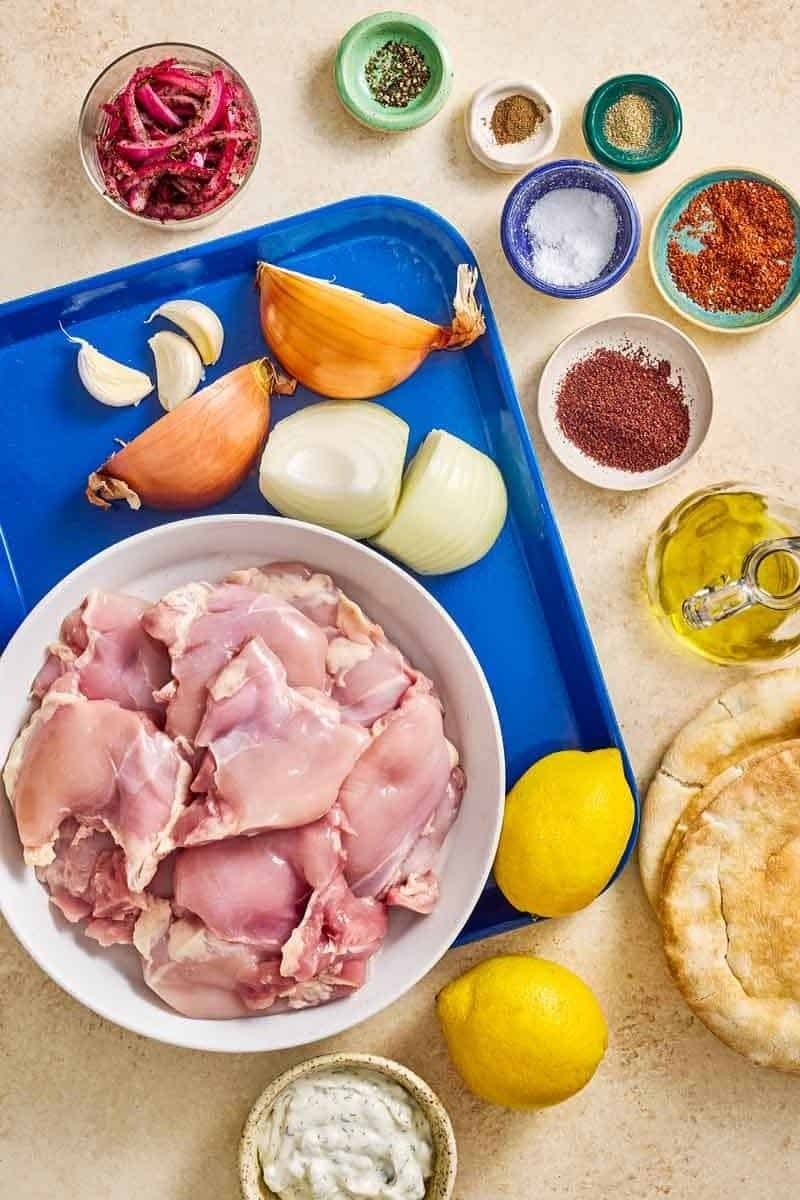 Raw chicken pieces in a bowl with garlic, onions, lemons, spices, olive oil, and pita bread arranged on a tray and table