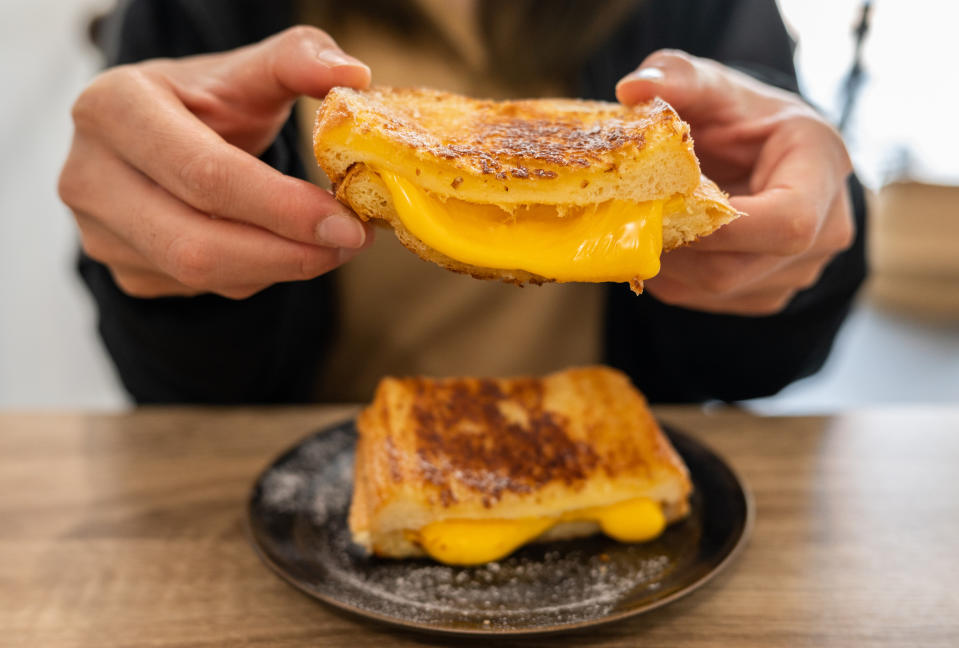 Person holding a grilled cheese sandwich with melted cheese stretching between two halves, over a wooden table with another sandwich on a black plate