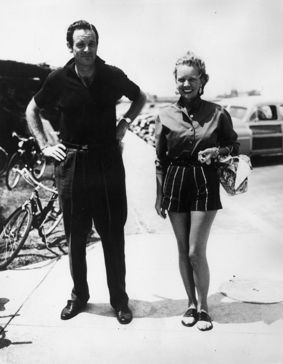 william woodward jr and ann woodward smile while standing outside on a sidewalk, he wear a polo shirt and slacks, she wears a collared button up shirt with striped shorts